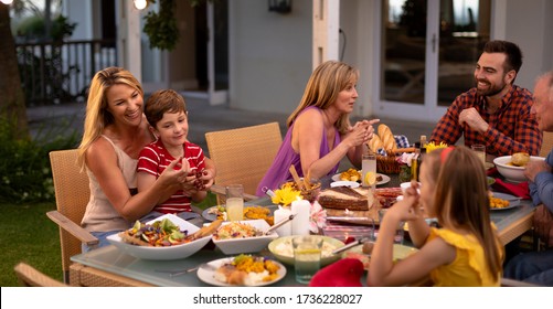 Side View Of A Multi-generation Caucasian Family Outside At A Dinner Table For A Celebration Meal, Eating And Talking, The Young Son Sitting On The Knee Of His Mother