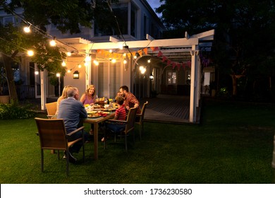 Vue latérale d'une famille caucasienne multi-génération assise dans le jardin devant leur maison à une table le soir pour un repas de fête ensemble, parler et manger