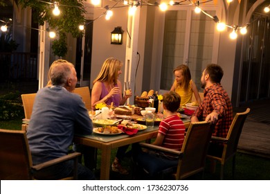 Side View Of A Multi-generation Caucasian Family Sitting Outside At A Dinner Table In The Evening For A Celebration Meal Together, Talking And Eating