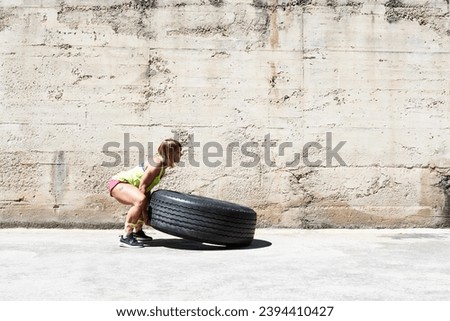 Side view of muscular woman lifting tire on sunny day outdoors. Cement textured wall in background. Copyspace.
