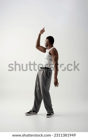 Side view of muscular young african american man in sleeveless t-shirt and pants looking away on grey background, contemporary shoot featuring casual attire