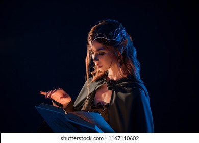Side view of mystic female beatiful elf reading magical book in darkness. Girl having long vawy hair, pretty face features, plump lips, big eyes, wearing cape on shoulders, elegant, thin crown.