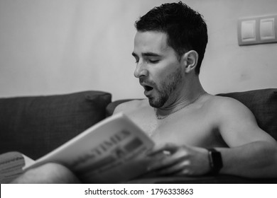 Side view of naked young man sitting on sofa and reading newspaper and yawning. Monochrome