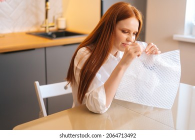 Side view of nervous young woman playing pops bubble wrap to calm herself sitting at table in kitchen. Stressed pretty redhead female popping plastic bubblewrap. Concept of mental health.