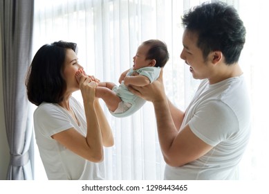 Side View Of New Family Activity Playing Together. Father Holding Newborn Baby Body While Mother Holding Legs And Kiss The Laughing Baby At Home. Happy Family Activity Concept.