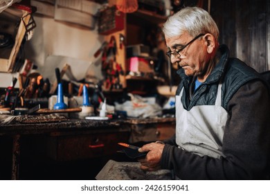 Side view of an old artisan sitting at his cobbler's workshop and cutting leather parts with scissors and preparing it for shoe making. A senior shoemaker is cutting a leather parts at cobbler's shop. - Powered by Shutterstock