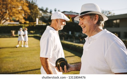 Side View Of An Old Man Standing In A Park Playing Boules On A Sunny Day. Senior Man In Hat Playing Boules In A Lawn With Friends.