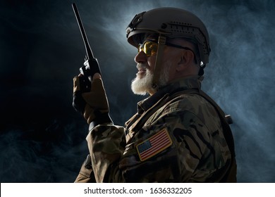 Side View Of Old Military Soldier In Uniform Holding Portable Radio Station In Smoke. Close Up Of Male Commander In Camouflage Uniform Giving Orders In Dark Atmosphere. Concept Of Army, Military.