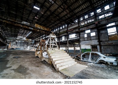 Side View Of An Old Rusty Antique Car Frame On The Background Of Broken-down Plant. Interior Of The Large Abandoned Building With A Damaged Car