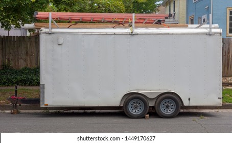 Side View Of Older White Utility Work Trailer Parked In Residential Neighborhood With Ladders On Top.