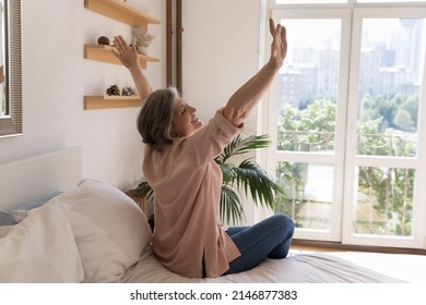 Side view older woman sit on bed in modern light warm bedroom raise hands stretch body muscles feels happy, welcomes new day, enjoy carefree morning, untroubled retired life and comfort living concept - Shutterstock ID 2146877383