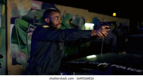 Side View On African American Young Cop Pointing Gun At Somebody Outdoors At Night. Police Officer Arresting Somebody At Dark Street At His Car.