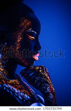 side view on awesome african model in neon light, unusual interesting portrait of beautiful girl with fluorescent makeup on whole body. isolated blue background