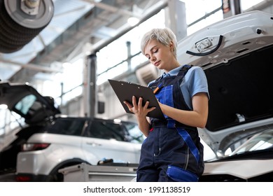 Side view on caucasian female Auto mechanic checking car engine, writing checklist on clipboard. lady in overalls uniform looking confident and serious, opened hood of car in the background