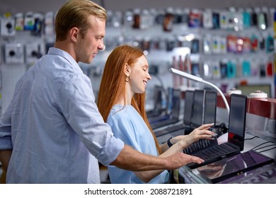 Side View On Couple Buying Laptop In Store, Choosing The Best Laptop For Work And Education. Caucasian Redhead Female And Handsome Guy In Casual Bright Blue Outfit Walking In Shop, Store