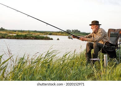Side view on elderly gray-haired man fishing on lake in nature, patience and recreation concept.Portrait of senior caucasian fisherman with rod sitting on chair in nature, countryside