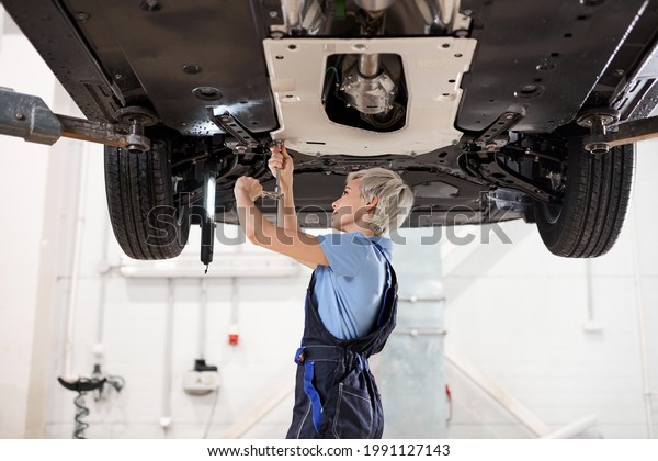 Side view on Female Auto Mechanic Working Underneath\
Car In Garage. Portrait of hardworking young short haired female\
mechanic in overalls, inspecting lifted car. Mechanic woman working\
in car shop