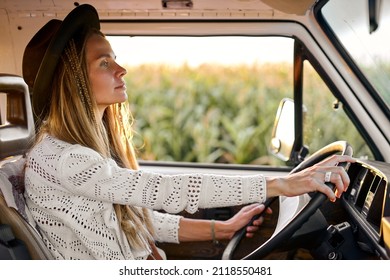 Side View On Focused European Caucasian Female In Hat With Long Hair Driving Mini Van, Through The Cornfield, Attractive Lady Exploring New Placed, Travelling, Hippie Lifestyle, Travel Concept.