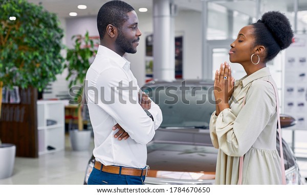 side view on\
good-looking cute african female begging her husband in car\
showroom, woman want to get new\
auto