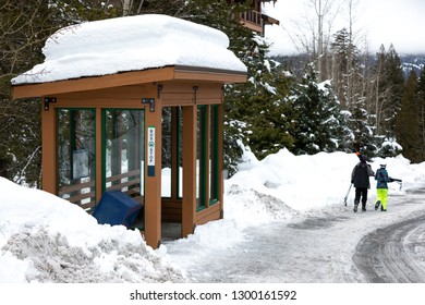 Side View On A Mountain Bus Stop And Wood Shelter Covered In Snow, With Skiers Walking Away In The Background