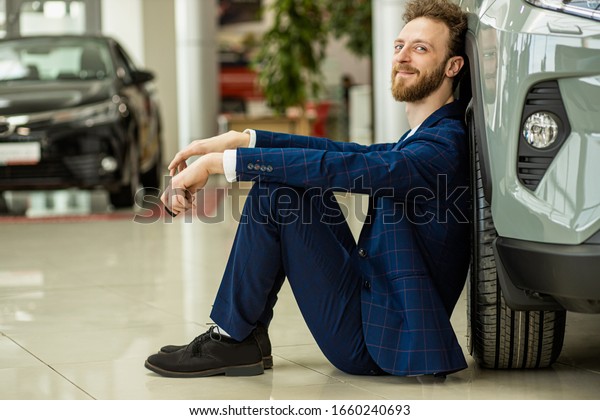 side view on positive smiling happy man buyer in\
dealership, man sit next to his new car, wearing formal suit, look\
at camera