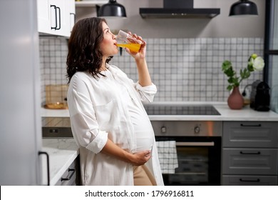 side view on pregnant woman drinking orange juice at home, young caucasian female enjoy time at home, hold hand on belly. in the kitchen - Powered by Shutterstock