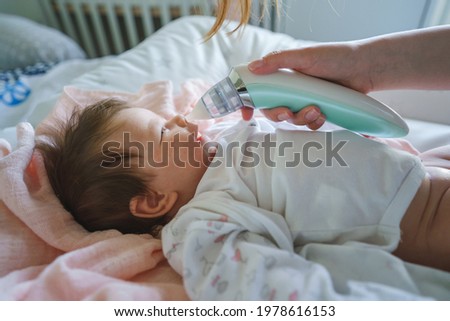 Side view on unknown woman mother using electric baby nasal aspirator mucus nose suction sucking the saliva from baby's nose cleaning while lying on the bed