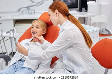 Side view on woman dentist in uniform works with child patient in dental office, showing patient's teeth on mirror, confidently explaining how to treat and which tooth will be after treatment - Shutterstock ID 2110592864