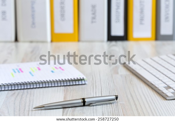 Side view on the working place\
of manager.\
Light wooden desk with color pen, notepad with marked\
handwriting, computer keyboard and line of folders on the\
background