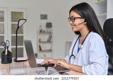 Side View On Young Asian Female Doctor In White Medical Lab Working On Laptop Computer Communicating Online With Patient. Primary Care Consultations, Psychotherapy, Emergency Services