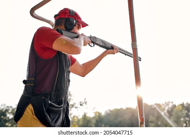 Side View On Young Caucasian Man Wear Ear Plug Confidently Aiming Shotgun At Target In Outdoor Shooting Range, Alone. Man Practicing Fire Weapon Shooting. Hobby, Skill, Shooting Concept.