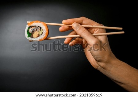 side view of one appetizing Philadelphia roll with salmon, eel, avocado, cucumber, green calve and cream cheese in hand holding with bamboo chopsticks on dark background