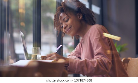 Side view of one happy young African woman writing indoor, Pretty Black college student study with laptop by the window doing homework writing letter