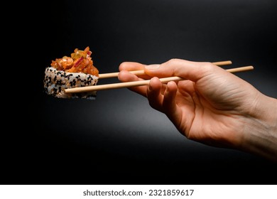 side view of one piece of roll with caramelized onion, raw fish, sesame and rice in hand holding with wooden chopsticks.