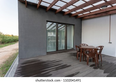 Side view of an open veranda in front of a modern forest cottage. Pine young forest under the sunset rays in the background.