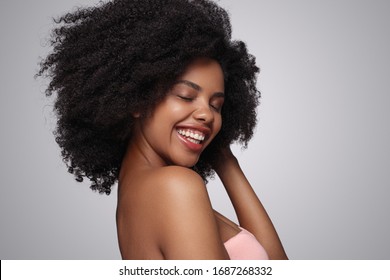 Seitenansicht der optimistischen Afroamerikanerin, die mit geschlossenen Augen lächelt und sauberes, lockiges Haar berührt, während sie den Spa-Service auf grauem Hintergrund darstellt