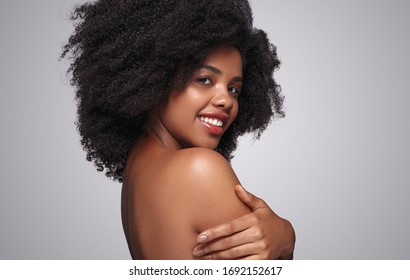 Side view of optimistic African American female smiling for camera and touching shoulder after spa procedure against gray background