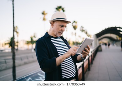 Side View Of Optimistic Elderly Male In Casual Outfit Standing Near Banister In Park While Walking Around City Using Tablet