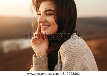 Side view of optimistic smiling millennial female with long dark hair looking away romantically while relaxing in sunlight in autumn evening in countryside