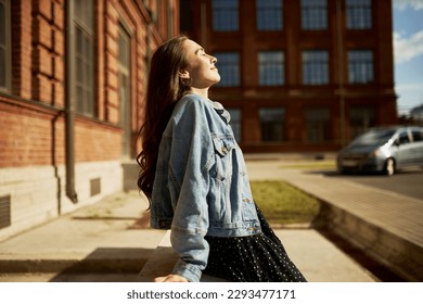 Side view outdoor image of gorgeous student girl with closed eyes in denim outfit leaning on edging at city streets taking sun bathing with face turned to sunlight, enjoying warm day and freedom - Shutterstock ID 2293477171