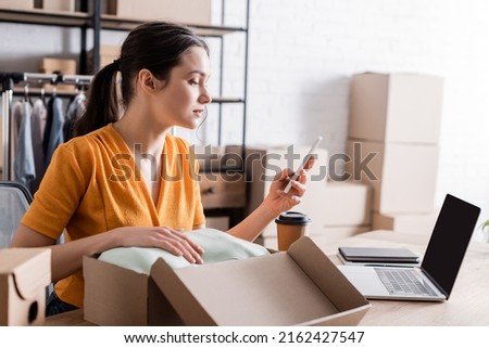 Side view of owner using smartphone near clothes in carton box in online web store