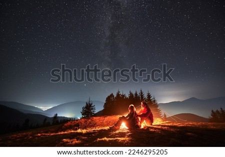 Side view of pair sitting one behind other on meadow near bright flame of bonfire and watching surrounding beauty of evening starry sky. Mountain hills and lighted hamlets in the distance.