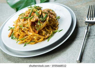 Side View Of Pan Asian Ramen Egg Noodles With Meat And Vegetables Served On Plate With Spring Onion Scallions, Sesame Seeds On Top. Steel Fork Near Glass Noodles Dish. Gray Stone Table Background