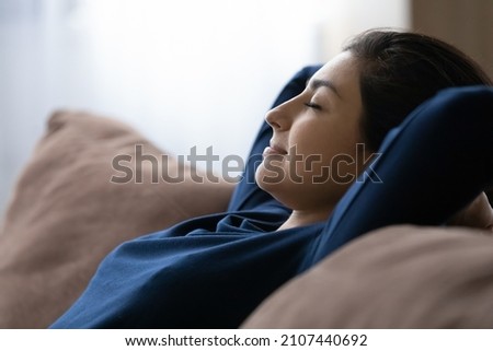 Side view peaceful happy young Indian woman resting on cozy sofa with closed eyes, daydreaming napping sleeping meditating breathing fresh air, enjoying carefree mindful leisure pastime.