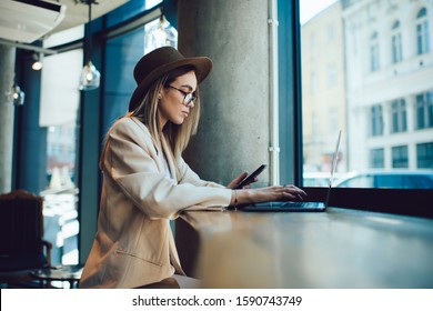 Side view of pensive elegant adult lady in glasses and hat holding phone and using laptop sitting at table near window in modern cafe