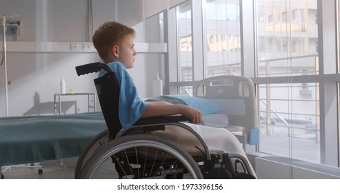 Side view of pensive kid sitting on wheelchair looking out of window in hospital ward. Unhappy sick preteen boy in wheelchair sitting alone in hospital room