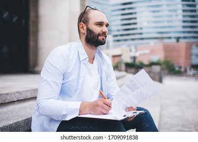 Side view pensive modern man in light shirt   jeans having break stairs   making plan paper blurred background 