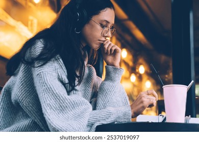 Side view of pensive young woman in headphones touching lips while browsing digital tablet in cafe of New York city - Powered by Shutterstock