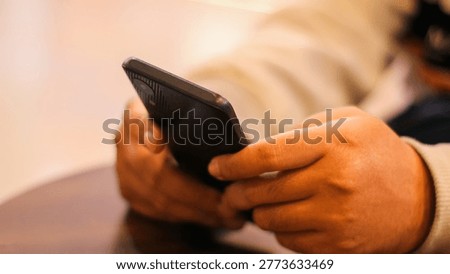 Side view of a person's hands holding and using a black smartphone while sitting at a table in a cafe.