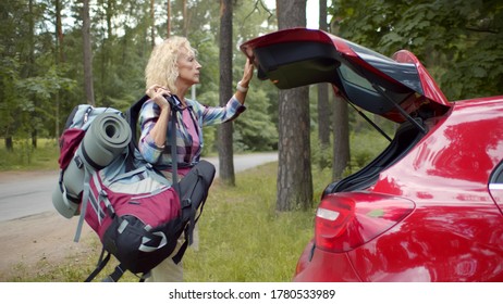 Side View Pf Attractive Mature Woman Loading Hiking Backpack In Red Car In Forest. Senior Female Traveler Packing Auto For Trekking Trip. Active Retirement And Tourism Concept